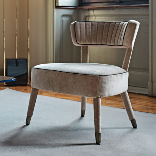 A Modern Velvet Occasional Armchair with four tapered legs sits on a light gray rug in a room with wooden floors and large windows. The plush beige chair features a cushioned seat and a horizontal-striped, upholstered backrest. The room boasts sophisticated, minimalist decor.