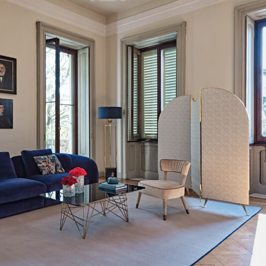 A sophisticated living room featuring a blue velvet sofa, a modern geometric glass coffee table adorned with red flowers, and the elegant touch of a Modern Velvet Occasional Armchair. A stylish floor lamp stands by the tall, wooded windows, and a decorative room divider completes the elegant ambiance.