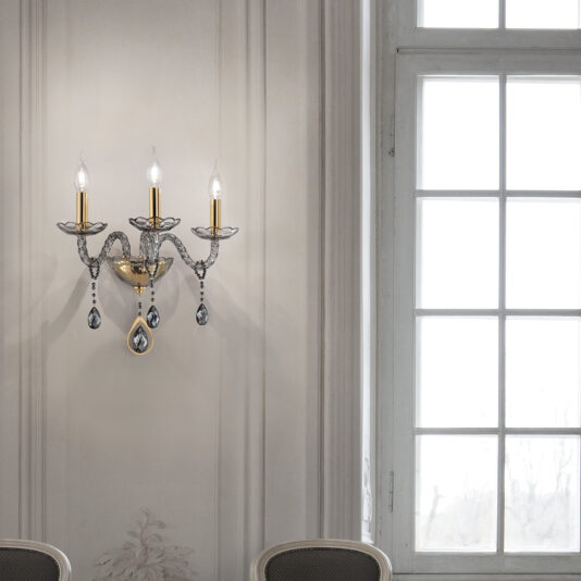 Smoked Glass 3 Arm Wall Light A Smoked Glass 3 Arm Wall Light, featuring three candle-shaped bulbs and adorned with crystal embellishments, is elegantly mounted on an ornate wall panel. The sconce is placed next to a large multi-paned window that floods the tastefully decorated room with natural light.