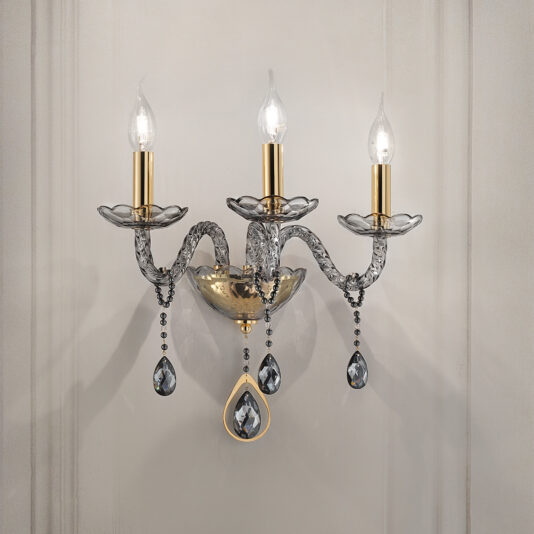 The Smoked Glass 3 Arm Wall Light is a decorative fixture featuring candle-shaped bulbs adorned with crystal-like accents hanging below each one. This sconce showcases elegantly curved arms, a textured central base, and boasts a gold and silver finish, all mounted on a light-colored wall.
