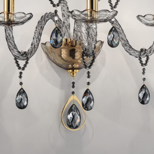 Smoked Glass 3 Arm Wall Light A close-up of the Smoked Glass 3 Arm Wall Light reveals its intricate design featuring curved smoked glass arms and crystal teardrop ornaments dangling from beaded chains. The central column, finished in polished gold, adds a touch of elegance to the overall look. This exquisite wall light is mounted on a light gray wall.