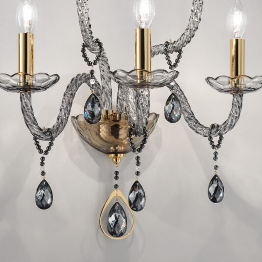 Smoked Glass 5 Arm Wall Light A close-up of an elegant chandelier featuring three light bulbs with gold-colored sockets. The arms of the chandelier are adorned with twisting clear glass, reminiscent of the Smoked Glass 5 Arm Wall Light, and hanging black crystal teardrops, adding a touch of sophistication and glamour to the design.