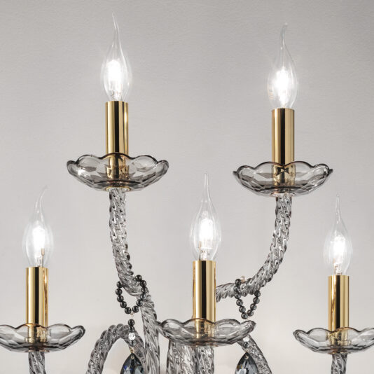 Smoked Glass 5 Arm Wall Light A close-up view of the Smoked Glass 5 Arm Wall Light featuring candle-shaped bulbs. The bulbs are mounted on gold-colored holders, which are supported by transparent, twisted glass arms adorned with ornate crystal accents. The background is a plain white wall.