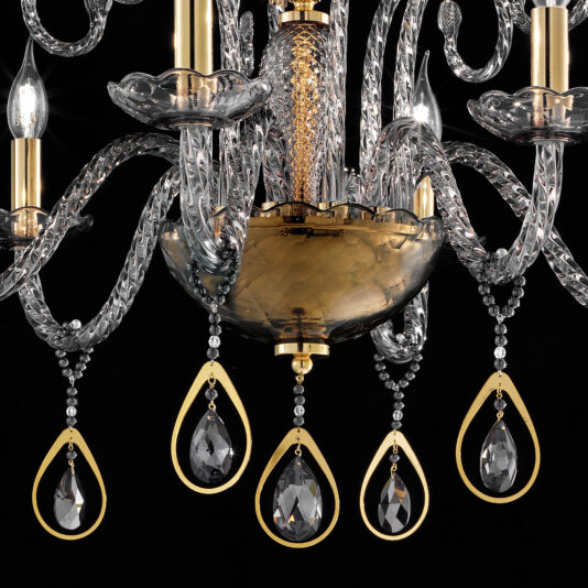 A close-up of the Smoked Glass Chandelier With Swarovski® Crystals And Pearls showcases its decorative elegance. It features multiple ornate arms with intricate curling designs, candle-shaped light bulbs, and dangling crystal pendants, all set against a black background. The central piece is finished in gold.