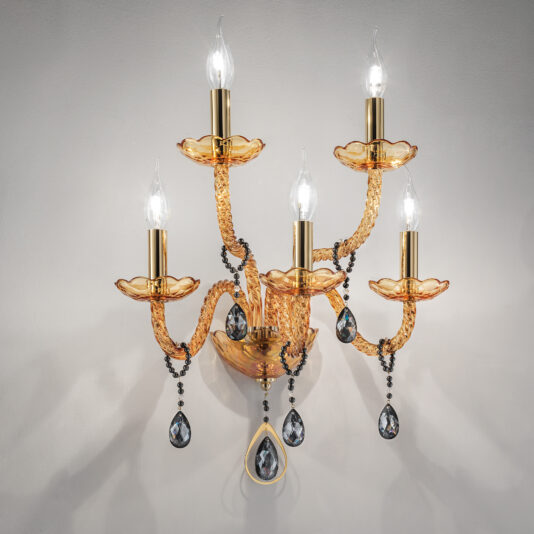 The Amber Glass 5 Arm Wall Light features a golden ornate design with five candle-shaped bulbs. This sconce, adorned with intricate, twisted arms and dangling black crystal-like ornaments, showcases beautiful amber glass accents. Its elaborate design stands out magnificently against the plain, light-colored wall background.