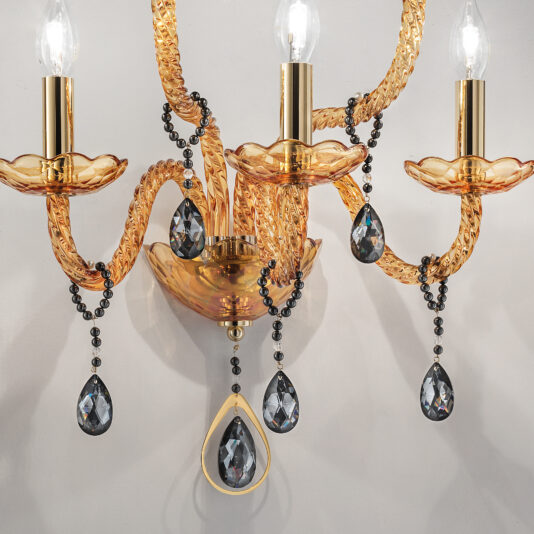 Amber Glass 5 Arm Wall Light A close-up view of the Amber Glass 5 Arm Wall Light with lit bulbs showcases each arm adorned with golden, braided designs and glass-like plates supporting the bulbs. Elegantly hanging from the fixture, black beaded chains embellished with large teardrop crystal pendants enhance its decorative appeal.