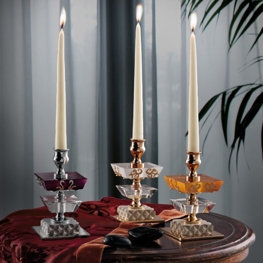 Three Elegant Coloured Glass Candlesticks, ornamented with intricate metal accents, stand on a table draped with a red cloth. Each candlestick supports a tall, lit, white taper candle. In the background, sheer curtains and the silhouette of a plant are visible.