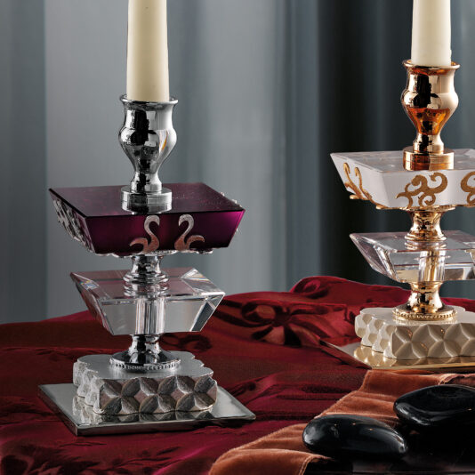Elegant Coloured Glass Candlestick A close-up of two Elegant Coloured Glass Candlesticks on a red and orange draped surface. Each holder features geometric and elaborate designs, with one showcasing a deep purple section and the other adorned with gold accents. A black stone decor item is visible in front.