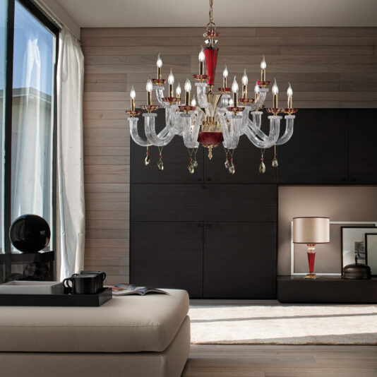 A modern living room boasts the Elegant Ruby Chandelier With Pendant Drops, adorned with crystal accents, suspended from the ceiling. A beige ottoman hosts a black teapot and matching teacup, while a black lamp with a red base stands beside it. A large window is dressed in sheer curtains completing the sophisticated look.