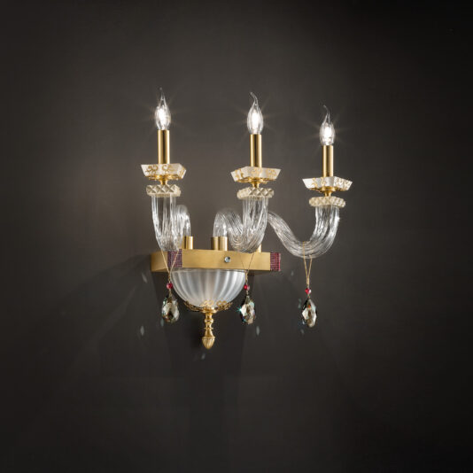 The Elegant Triple Wall Light With Pendant Drops is a gold sconce featuring intricate glass detailing and dripping crystal ornaments. Each arm holds a candle-shaped bulb that casts a warm glow against a dark background.