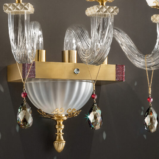 The Elegant Triple Wall Light With Pendant Drops brings a touch of luxury with its clear, curved glass arms complemented by gold accents and a frosted glass bowl. Adorned with hanging faceted crystal pendants and red beads, this sophisticated fixture casts intricate reflections against a dark backdrop.