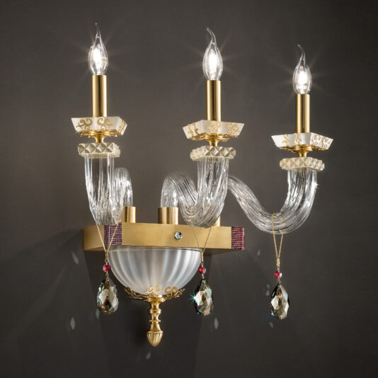 The Elegant Triple Wall Light With Pendant Drops is a sophisticated wall-mounted sconce featuring three candle-shaped bulbs. This ornate gold and glass design includes intricate scrollwork, crystal accents, and hanging teardrop-shaped crystals that surround a central frosted glass bowl, providing an exquisite look against a dark matte wall.