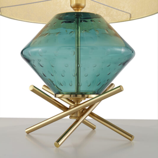 A close-up view of the Hand Blown Glass Table Lamp reveals a teal, diamond-shaped base adorned with bubble details. The lamp is supported by a brass stand featuring three intersecting rods, and it is topped with a partially visible beige lampshade. The overall design exudes modern elegance.