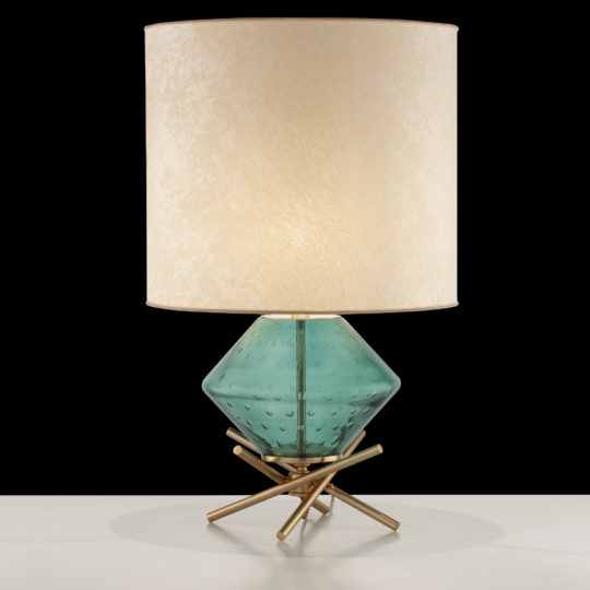 The Hand Blown Glass Table Lamp features a modern design with a cylindrical beige lampshade atop a blue-green diamond-patterned glass base. The base is supported by a geometric, gold-colored metal stand and is showcased on a white surface against a black background.