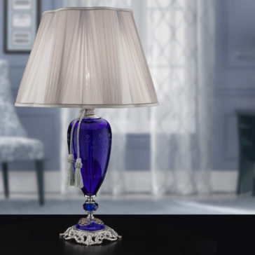 The Large Blue Hand-Blown Glass Table Lamp boasts a stylish design with a vibrant base adorned with decorative tassels. It features a classic white fabric lampshade and stands on an ornate metallic stand. This elegant lamp is set in a softly lit, elegantly decorated room with blurred background details.