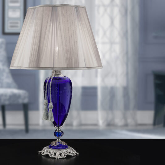 The Large Blue Hand-Blown Glass Table Lamp boasts a stylish design with a vibrant base adorned with decorative tassels. It features a classic white fabric lampshade and stands on an ornate metallic stand. This elegant lamp is set in a softly lit, elegantly decorated room with blurred background details.