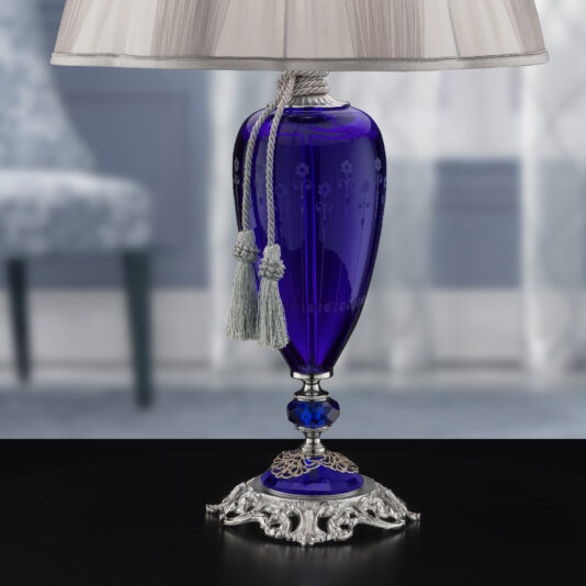 The Large Blue Hand-Blown Glass Table Lamp boasts a sophisticated design with a deep blue glass base adorned with white rope and tassels, enhanced by intricate silver detailing. The light-colored lampshade has subtle pleats, set against a softly blurred background of blue and gray hues.