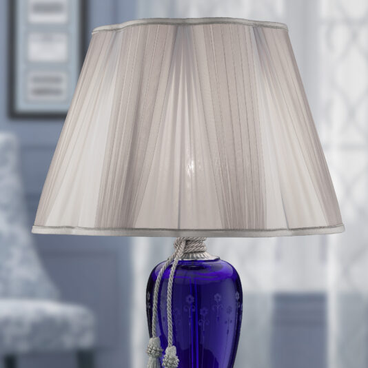 A Large Blue Hand-Blown Glass Table Lamp with a pleated white lampshade sits on a table. The base is adorned with a decorative white cord and tassels. The background is softly blurred, showcasing a light-colored room with subtle furnishings.