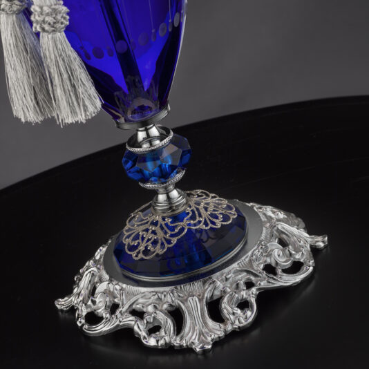 A close-up of the Large Blue Hand-Blown Glass Table Lamp showcases its elaborate silver base, a dark blue glass section with intricate detailing, and delicate light-colored tassels at the top. The dark background accentuates the lamp's elegance.