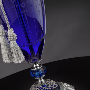 Close-up of the Large Blue Hand-Blown Glass Table Lamp featuring intricate silver detailing at the base and a few light grey tassels hanging from its neck. The dark background highlights the fine craftsmanship and rich color of the glass.