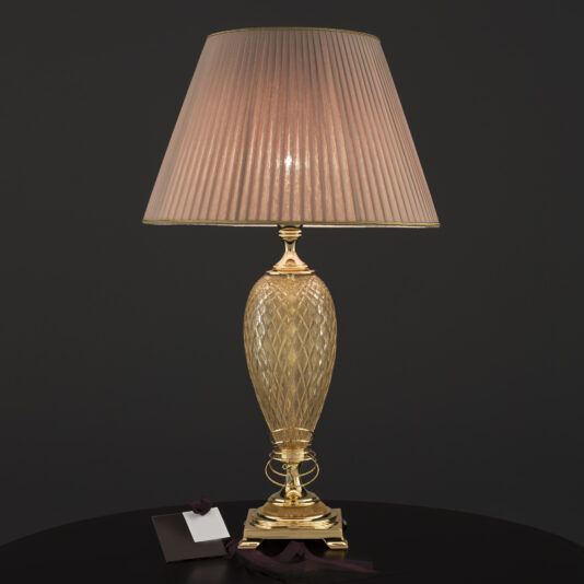 A Large Glass Table Lamp With Eternity Charm, featuring a patterned gold-colored base and a pleated beige lampshade, sits on a black round table. A small white card with a dark ribbon lies next to the lamp. The background is a solid dark color.