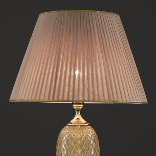 Large Glass Table Lamp With Eternity Charm The Large Glass Table Lamp With Eternity Charm stands elegantly, featuring a pleated beige lampshade and an ornate base with a diamond pattern. The lamp casts a warm, soft light against a dark background, highlighting its eternity charm design.