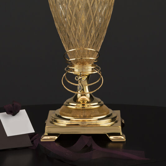 Large Glass Table Lamp With Eternity Charm A gold-colored trophy with an intricate diamond-patterned design sits on a square base adorned with ribbon-like decorations. Nearby, beside the Large Glass Table Lamp With Eternity Charm, is a black card with a white insert tied with a dark purple ribbon. The background is solid black.