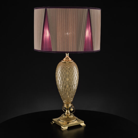 The Large Glass Table Lamp With Love Charm features an ornate gold-colored base and a pleated lampshade. The shade boasts a light brown exterior with subtle red and pink tones that are visible when illuminated. Adorned with a delicate love charm, the lamp is placed on a dark surface against a black background.