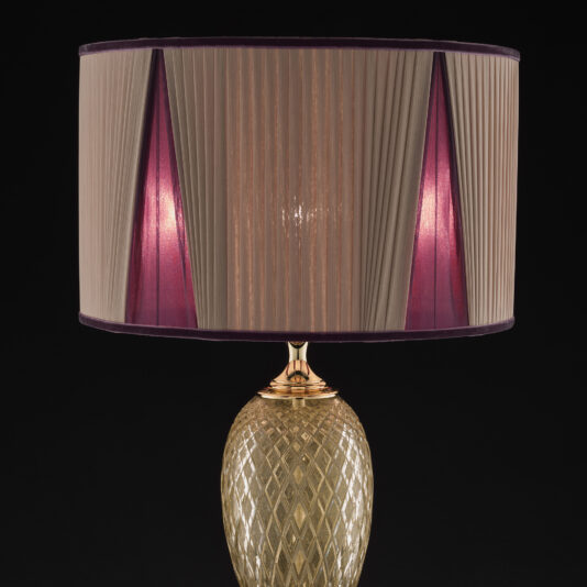 Introducing the Large Glass Table Lamp With Love Charm: a stylish table lamp featuring a gold, diamond-patterned base and a cylindrical, pleated lampshade with alternating beige and pink segments. This elegant and modern design is adorned with a love charm and set against a striking black background.