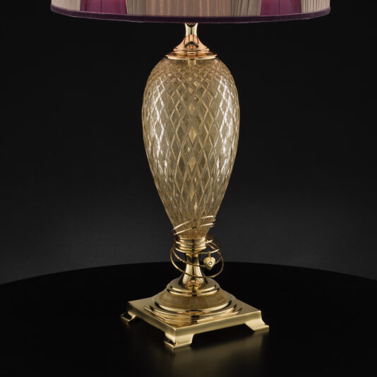 The Large Glass Table Lamp with Love Charm features a lustrous gold, diamond-patterned base and a sleek, metallic gold stand. It includes a fabric shade in a deep purple hue with an elegant striped pattern against a dark backdrop. Adding to its charm, the lamp also comes adorned with a delicate love charm.