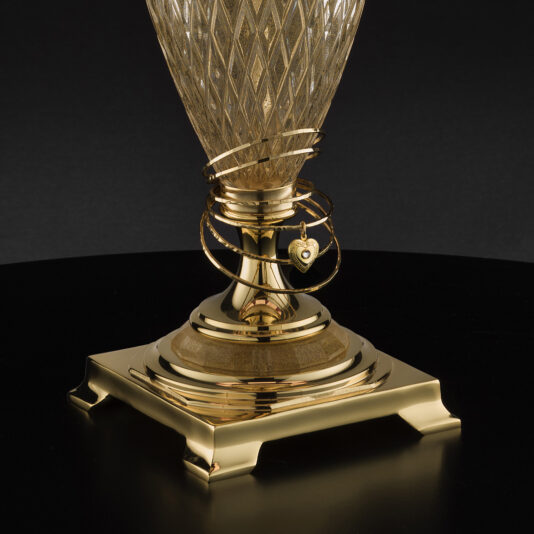 A close-up of the Large Glass Table Lamp With Love Charm, featuring an intricate diamond-patterned base. A gold bracelet, adorned with a small heart-shaped love charm, is elegantly draped around the lamp's base, which rests on a dark, reflective surface.