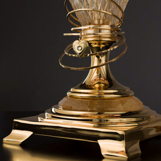 Here is a close-up view of the ornate base of the Large Glass Table Lamp With Love Charm, featuring intricate golden designs and swirls. Dangling around its neck is a heart-shaped locket with a white center, reminiscent of a love charm. The dark background accentuates the shiny golden surface of the lamp's base.