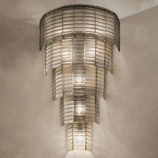 Large Tiered Murano Glass Wall Light The Large Tiered Murano Glass Wall Light, featuring a metallic sconce and rectangular translucent panels, emits a warm and diffused light. With its art deco style, the layered design creates a captivating cascading effect against a plain, neutral-colored wall.