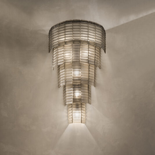 The Large Tiered Murano Glass Wall Light features a modern design with geometric tiers of overlapping metallic panels, emitting a soft, warm glow. The cascading arrangement creates intricate patterns of light and shadow on the textured wall behind it.