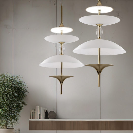Modern interior featuring three contemporary pendant lights, including the Large Modern Venetian Glass Pendant Light with white and brass finishes, hanging at different heights against a minimalist gray wall. A wooden shelf below holds a small arrangement of books, and a lush green potted plant adds a touch of nature.