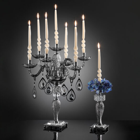 A luxurious Smoked Glass Candelabra Style Candlestick with eight lit candles and ornate details stands next to a classic candlestick adorned with blue flowers. Set against a dark background, the design is intricate, featuring reflective surfaces and hanging crystal adornments.