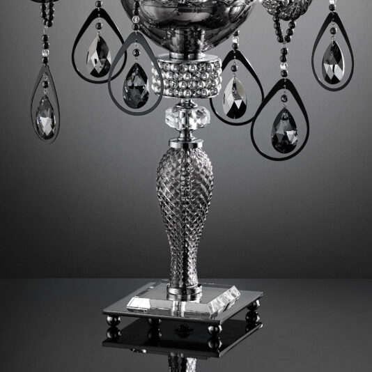 Smoked Glass Candelabra Style Candlestick A close-up of the **Smoked Glass Candelabra Style Candlestick** featuring an ornate, reflective design with a textured stem and decorative teardrop-shaped crystal ornaments adorning the top. Crafted from smoked glass, it is set against a smooth gradient background that transitions from dark gray to black, enhancing its brilliance.