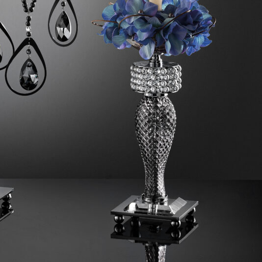 Smoked Glass Candelabra Style Candlestick A bouquet of vibrant blue and purple flowers is held in a shiny, silver, intricately designed vase adorned with pearl-like beads near the top. The vase gleams beside a Smoked Glass Candelabra Style Candlestick. In the background, partially visible hanging decorative black and clear crystal ornaments add to the scene's elegance. The surface is reflective.