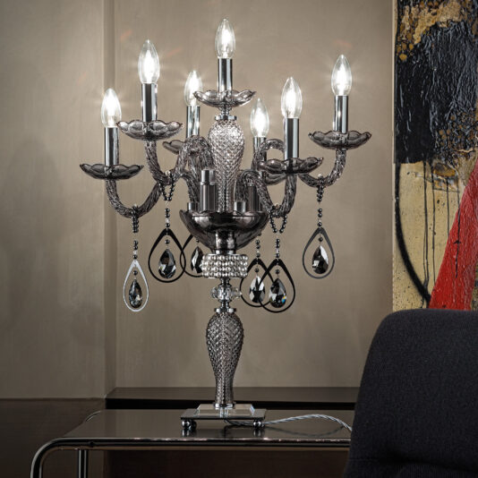 A Smoked Glass Candelabra Style Table Lamp with a black and silver ornate design and multiple bulbs is displayed on a modern table. Behind the lamp is a beige wall featuring part of an abstract painting in red and black hues. A dark chair is partially visible in the foreground.