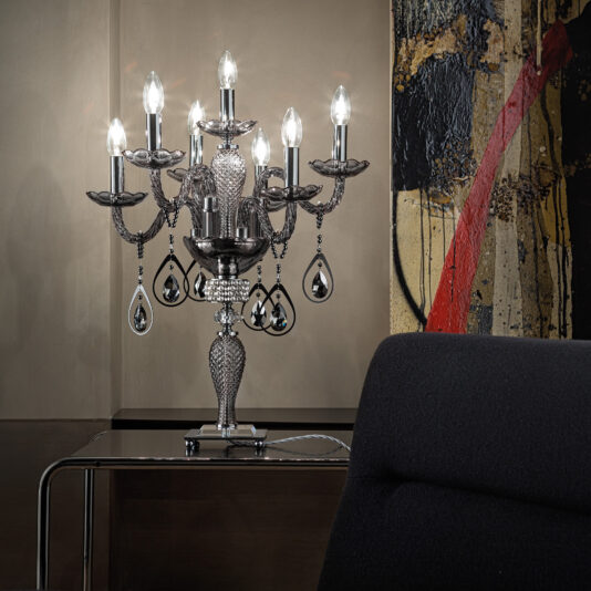 Smoked Glass Candelabra Style Table Lamp A **Smoked Glass Candelabra Style Table Lamp** with multiple crystal-adorned arms and lit bulbs sits on a polished table in a dimly-lit room. Its reflection casts a soft glow on the beige wall, reminiscent of its elegant design. Modern abstract artwork featuring bold red and yellow strokes is displayed in the background.