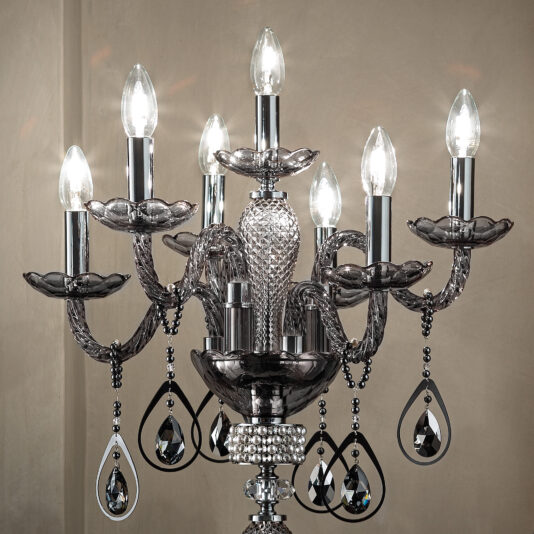 Smoked Glass Candelabra Style Table Lamp The Smoked Glass Candelabra Style Table Lamp is a sophisticated piece featuring seven lit candle-shaped bulbs on silver arms with intricate glass detailing. Reminiscent of an elegant, ornate chandelier, this lamp includes several crystal pendants hanging from the arms, enhancing its sparkle and sophistication against a neutral backdrop.