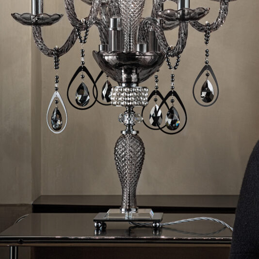 Smoked Glass Candelabra Style Table Lamp A close-up of an elegant black crystal chandelier with intricate detailing and multiple teardrop-shaped crystal pendants. The chandelier, resembling the Smoked Glass Candelabra Style Table Lamp, is mounted on a reflective surface, adding to the overall luxurious appearance.