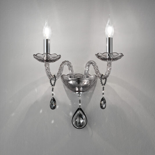 The Smoked Glass Double Wall Light is a stylish fixture with two illuminated bulbs, showcasing a sleek silver design. It features intricately curved arms, clear decorative elements, and dangling teardrop-shaped crystals, all set against a plain gray background.