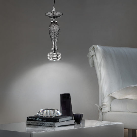 A sleek, modern living space showcases a Swarovski® Crystal And Pearl Pendant Spot Light suspended above a white side table. The table displays a stack of books, a crystal ashtray, a dark vase, and a candle. In the background, part of a white upholstered chair can be seen.