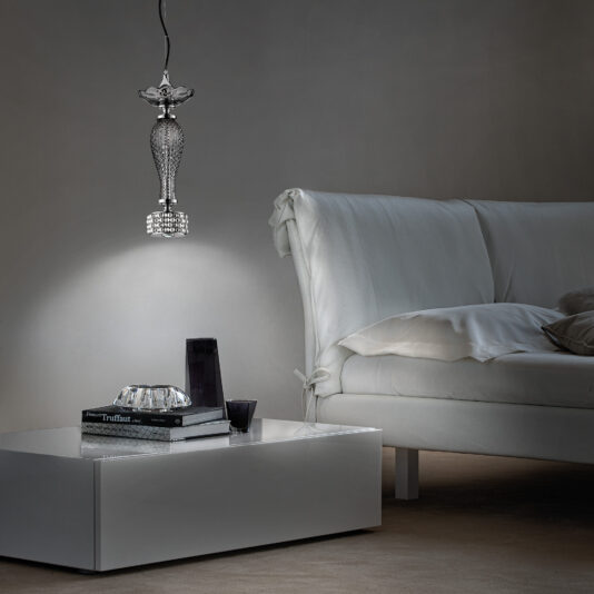A chic living room showcasing a white couch adorned with pillows, a sleek white coffee table displaying books and decorative glass accents, and an elegant Swarovski® Crystal And Pearl Pendant Spot Light illuminating the space from above. The room exudes a minimalist and sophisticated vibe, characterized by dim lighting and neutral hues.
