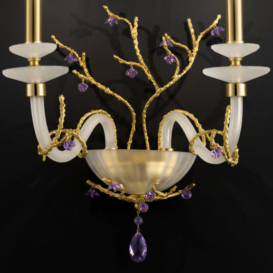 Candle Style Wall Light With Swarovski Crystal Flowers The Candle Style Wall Light With Swarovski Crystal Flowers is mounted against a black background, showcasing a decorative golden branch design. It features two frosted glass candle holders on either side, elegant curved glass arms, and a central bowl-like element adorned with additional purple accents.