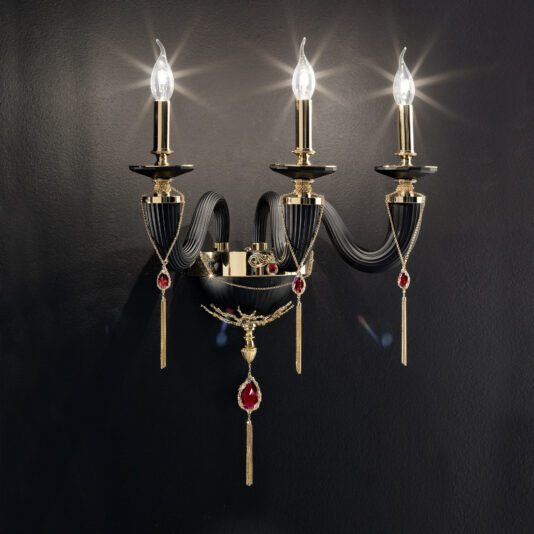 The Classic Black Triple Wall Light With Swarovski® Crystals is a luxurious wall-mounted gold candle-style light fixture featuring three flame-shaped bulbs. It is adorned with black and gold accents, red gemstone details, and ornate hanging elements, all of which stand out beautifully against a dark background.