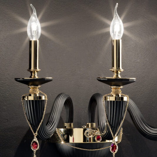 Classic Black Triple Wall Light With Swarovski® Crystals The Classic Black Triple Wall Light With Swarovski® Crystals features two luxurious, candle-shaped bulbs set in ornate gold holders. The design boasts fluted elements and draped gold chains adorned with red gemstones, all against a dark background.