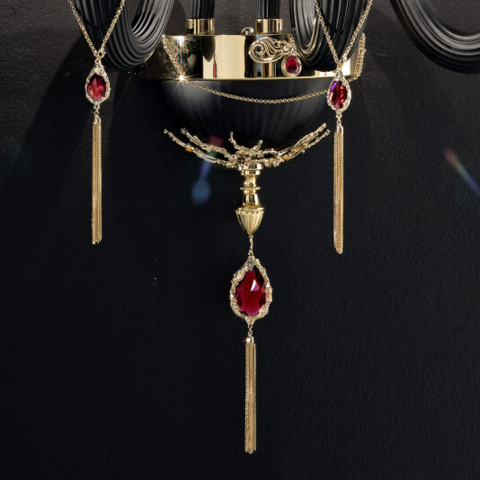 Classic Black Triple Wall Light With Swarovski® Crystals An ornate wall sconce with a gold base is adorned with red gemstones and intricate details. Elegant tassels hang below the main gemstone, creating a luxurious and sophisticated design. The Classic Black Triple Wall Light With Swarovski® Crystals elegantly contrasts against the dark background.