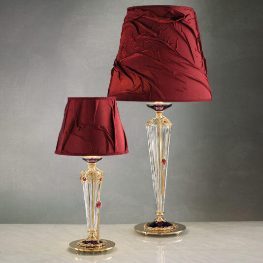 A pair of sophisticated Classic Style Swarovski® Crystal Table Lamps features red crinkled fabric shades and ornate gold accents that evoke timeless elegance. The clear, tapered glass bases are adorned with intricate gold and red details, mirroring the essence of genuine Swarovski® Crystal craftsmanship. The larger lamp is displayed behind and slightly to the right of its smaller counterpart.
